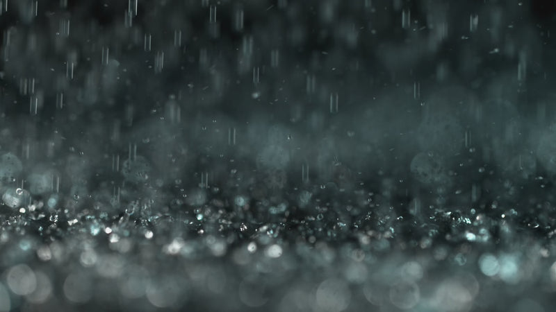 Close-up of soft rain falling in slow motion, with water droplets splashing gently on a dark surface — evoking calm, stillness, and deep relaxation.
